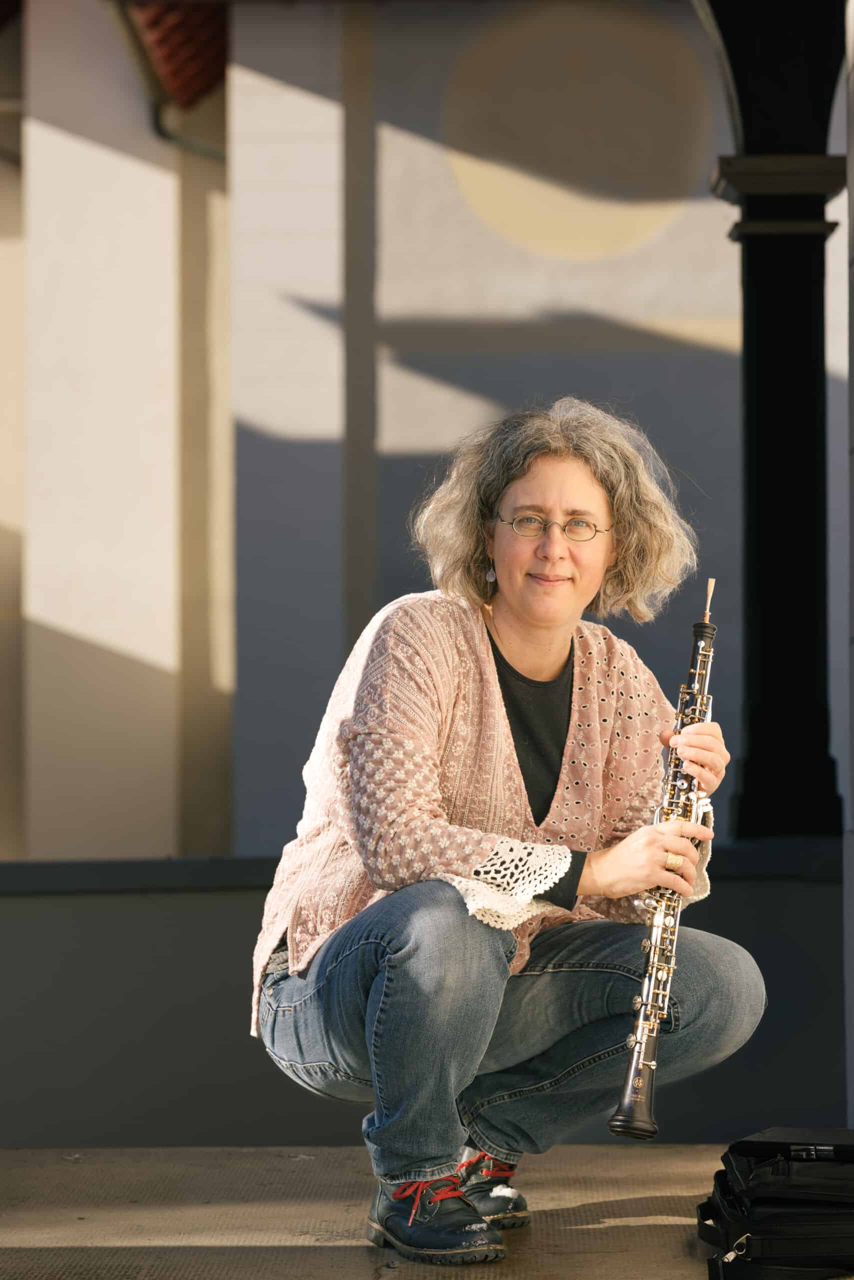 Eine Frau mit grauen Locken, Brille und einer rosa Spitzenstrickjacke hockt im Freien, hält eine Oboe in der Hand und lächelt leicht – ihre Haltung fängt einen stillen „Über mich“-Moment ein, während das Sonnenlicht geometrische Schatten an die Wand hinter ihr wirft. Eine Frau mit grauen Locken, Brille und einer rosa Spitzenstrickjacke hockt im Freien, hält eine Oboe in der Hand und lächelt leicht – ihre Haltung fängt einen stillen „Über mich“-Moment ein, während das Sonnenlicht geometrische Schatten an die Wand hinter ihr wirft.
