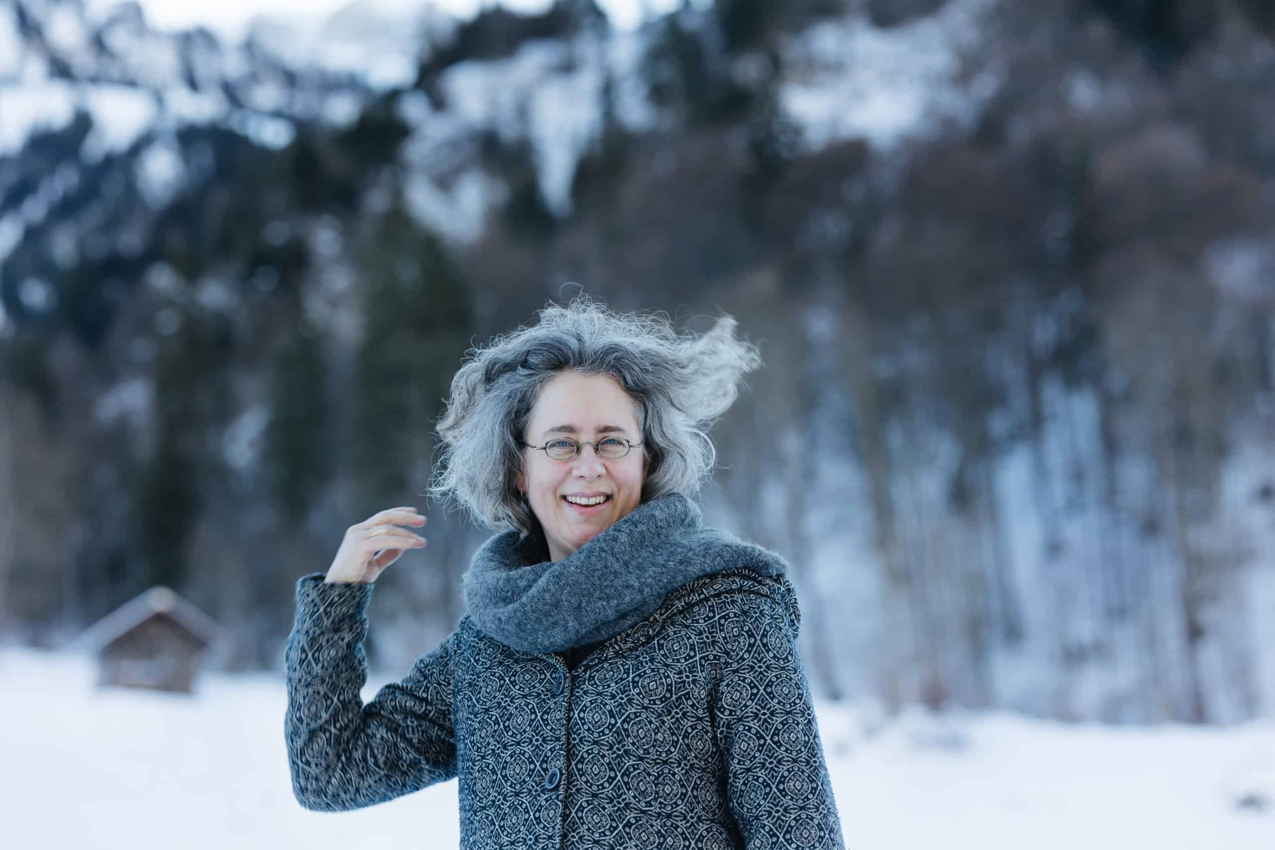 Eine Frau mit grauen Locken und Brille steht draußen in einer verschneiten Landschaft, bekleidet mit einem gemusterten Mantel und einem dicken Schal – ein authentischer „Über mich“-Moment, eingerahmt von verschwommenen Bäumen und fernen Bergen. Eine Frau mit grauen Locken und Brille steht draußen in einer verschneiten Landschaft, bekleidet mit einem gemusterten Mantel und einem dicken Schal – ein authentischer „Über mich“-Moment, eingerahmt von verschwommenen Bäumen und fernen Bergen.