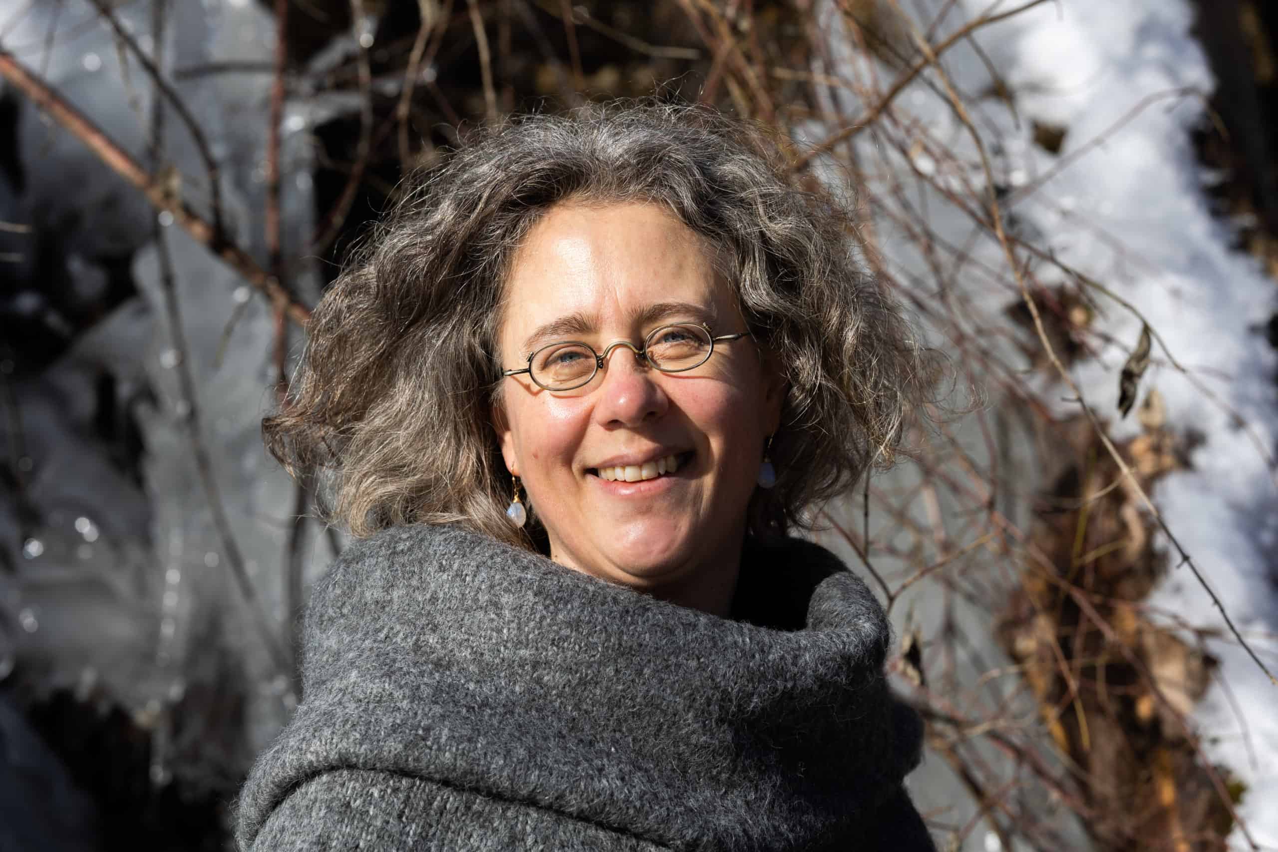 Eine Frau mit grauen Locken, Brille und einem grauen Schal lächelt draußen im Sonnenlicht – ein einladender „Über mich“-Moment, eingerahmt von kahlen Ästen und frischem Schnee. Eine Frau mit grauen Locken, Brille und einem grauen Schal lächelt draußen im Sonnenlicht – ein einladender „Über mich“-Moment, eingerahmt von kahlen Ästen und frischem Schnee.