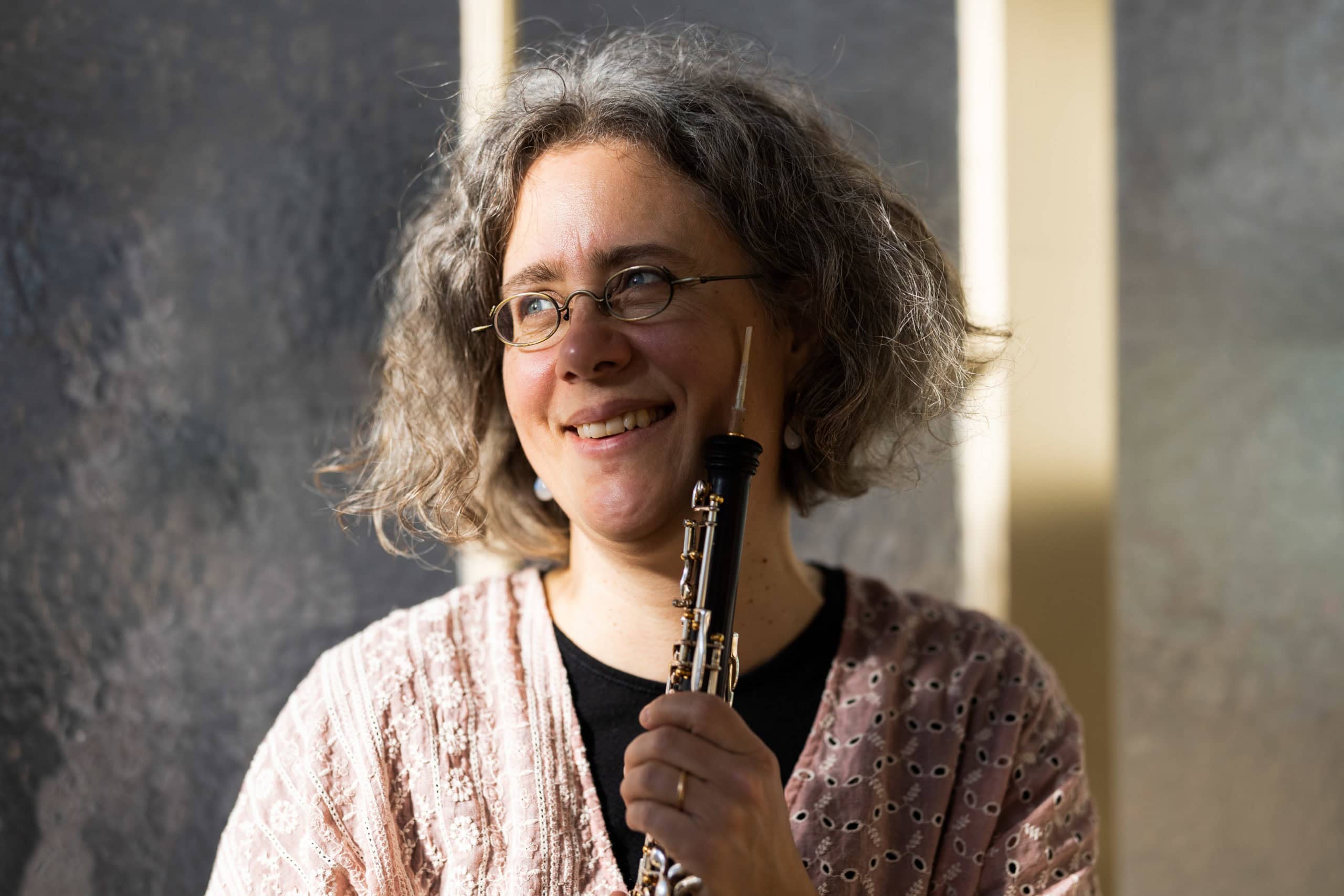 Eine Person mit grauen Locken und Brille lächelt, während sie eine Oboe aufrecht hält; sie trägt ein hellrosa gemustertes Oberteil und steht in sanftem, natürlichem Licht vor einem unscharfen Hintergrund – perfekt für eine „Über mich“-Vorstellung. Eine Person mit grauen Locken und Brille lächelt, während sie eine Oboe aufrecht hält; sie trägt ein hellrosa gemustertes Oberteil und steht in sanftem, natürlichem Licht vor einem unscharfen Hintergrund – perfekt für eine „Über mich“-Vorstellung.